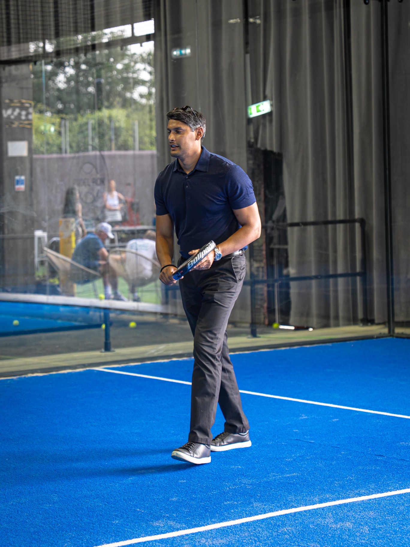 Guest in a navy polo walking onto the blue padel court with a racket
